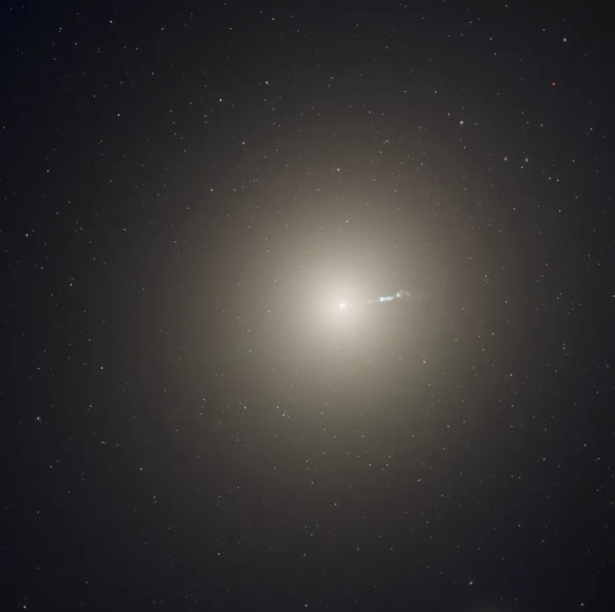 Messier 87 (M87), a giant elliptical galaxy with a bright core and a visible jet of energetic particles shooting out from its center, surrounded by a smooth, diffuse halo of stars, captured in fine detail against a dark space background.