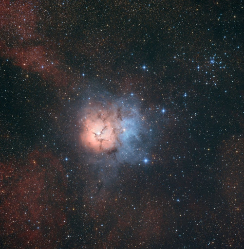 The Trifid Nebula - a distinct three-lobed structure. The image highlights the bright pink emission nebula, deep blue reflection nebula, and dark dust lanes, set against a dense star field.